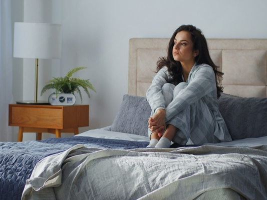 Woman sitting alone on a bed with her arms around her knees looking thoughtful in soft morning light