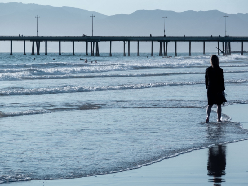 Woman standing alone at the shoreline with a pier in the distance and her reflection on the wet sand representing quiet reflection on emotional distance in a relationship