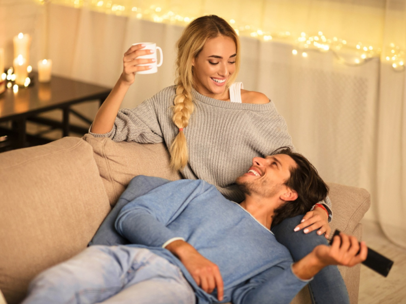 oung couple relaxed together on a couch with fairy lights and candles in the background representing an easy unpressured evening together